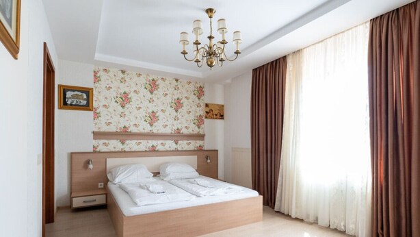 A bedroom with a wooden bed, floral wallpaper, a chandelier, and a large window with curtains. - FarawayHomes Aparthotel Vienna Airport #1 (Bruck an der Leitha)