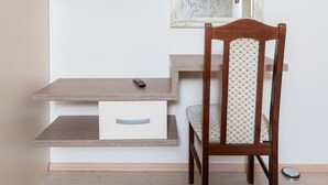 Una habitación con un televisor montado en la pared, una silla de madera, un escritorio pequeño con cajones y un espejo con marco decorativo.