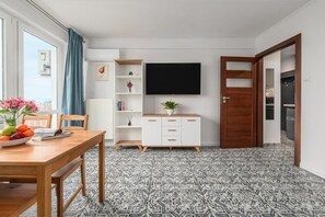 Ein modernes Wohnzimmer mit Fernseher, einem Holzes Esstisch mit Früchten, einem weißen Schrank und einem Fußboden mit Muster.