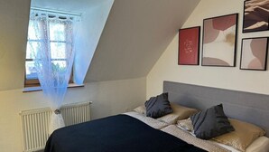 Une chambre à coucher avec un lit, un radiateur, une fenêtre avec des rideaux légers et des œuvres d’art encadrées accrochées au mur.