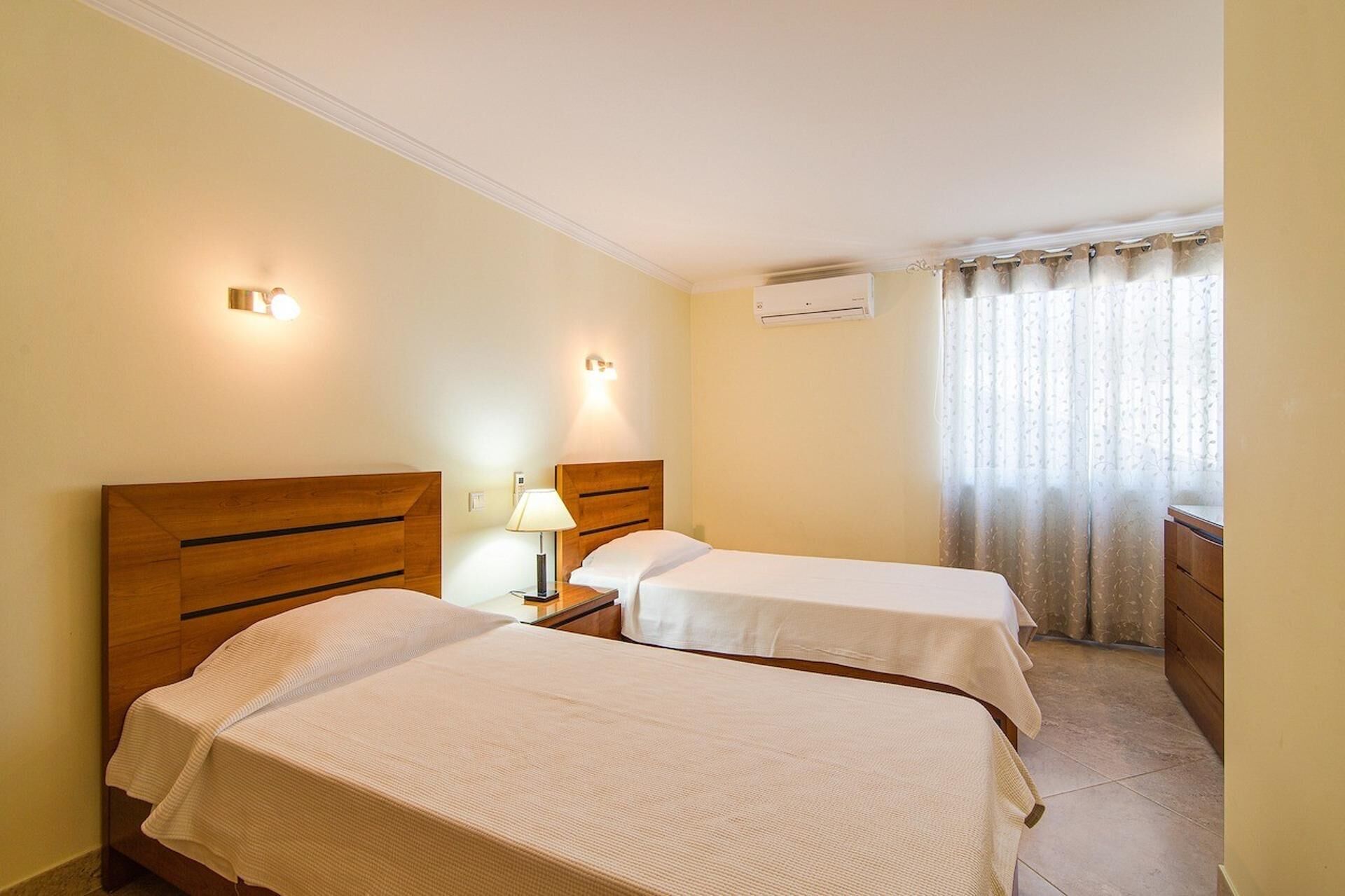 A hotel room with two beds, a nightstand with a lamp, an air conditioning unit, and a window with curtains.