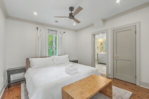 A bedroom with a bed, bedside tables, a wooden bench, a ceiling fan, and a door leading to a bathroom. - Charleston Charm: Renovated 4BR House with Modern Amenities (Charleston)