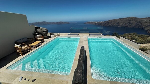 A pool area with two rectangular pools, a seating area with chairs and a table, and a view of the sea and hills.