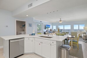 A modern kitchen with a dishwasher, white cabinets, and a dining area with a table and chairs. - Admiral's Quarters 1202 by Liquid Life (Orange Beach)
