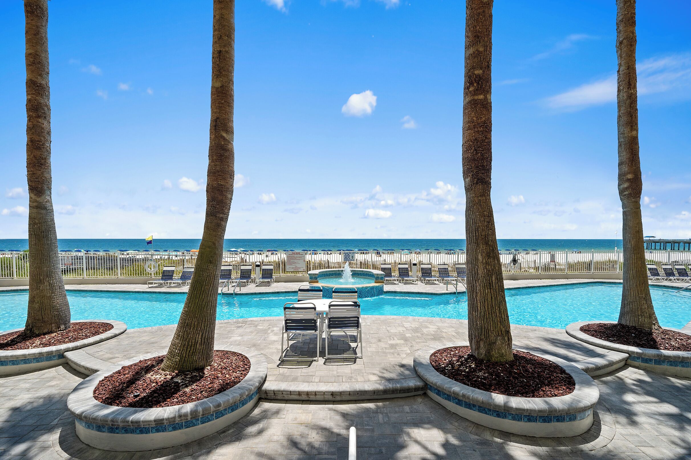 A pool area with lounge chairs, palm trees, and a view of the ocean.