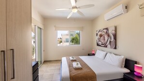 A hotel room with a bed, a ceiling fan, an air conditioner, a window with a view, and a painting on the wall. - Protaras Seaside Villa Andria (Paralimni)