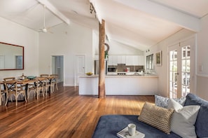 A spacious living area with a dining table, wooden flooring, a sofa with cushions, and a kitchen in the background. - PEACEFUL AND SERENE RURAL COTTAGE SITUATED IN QUINDALUP ON 4.5 ACRES (Quindalup)