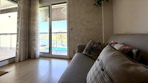 A living room with a sofa, a sliding glass door leading to a balcony, and a hanging plant. - Tranquil apartment, pets welcome - 4 guests (Roses)