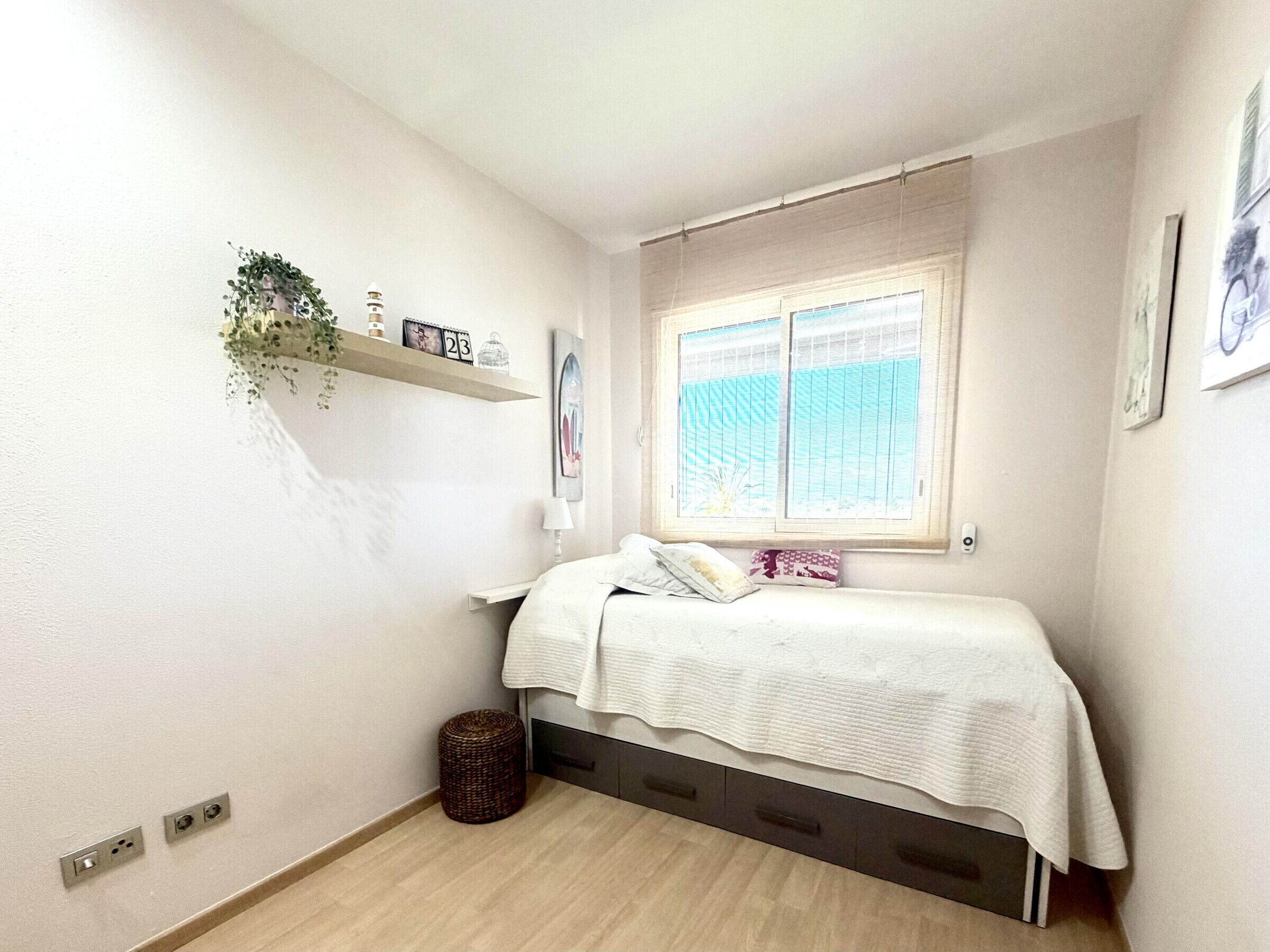 Une petite chambre à coucher avec un lit, une fenêtre avec des stores, une étagère avec des plantes et une horloge murale.