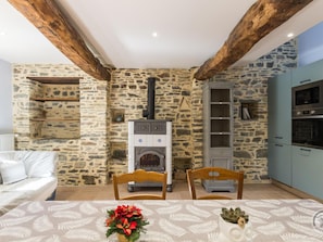 A stone-walled room with a fireplace, wooden beams, a dining table with chairs, and a kitchen area with cabinets and appliances. - Peaceful house, authentic setting - 6 guests (Bretteville-sur-Ay)