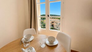 Un comedor con una mesa dispuesta para dos, una ventana con vistas a edificios y vegetación, y una puerta con manija.