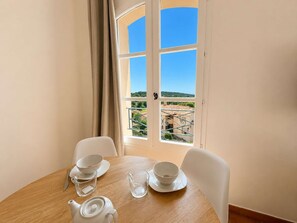 Ein Essbereich mit einem Tisch für zwei Personen, einem Fenster mit Blick auf Gebäude und Grünflächen sowie einer Tür mit Griff.