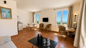 Una sala de estar con sofá, una mesa de comedor con sillas, televisor montado en la pared y ventanas amplias con cortinas.