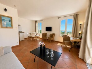 Ein Wohnzimmer mit einer Couch, einem Esstisch mit Stühlen, einem an der Wand befestigten Fernseher und großen Fenstern mit Vorhängen.