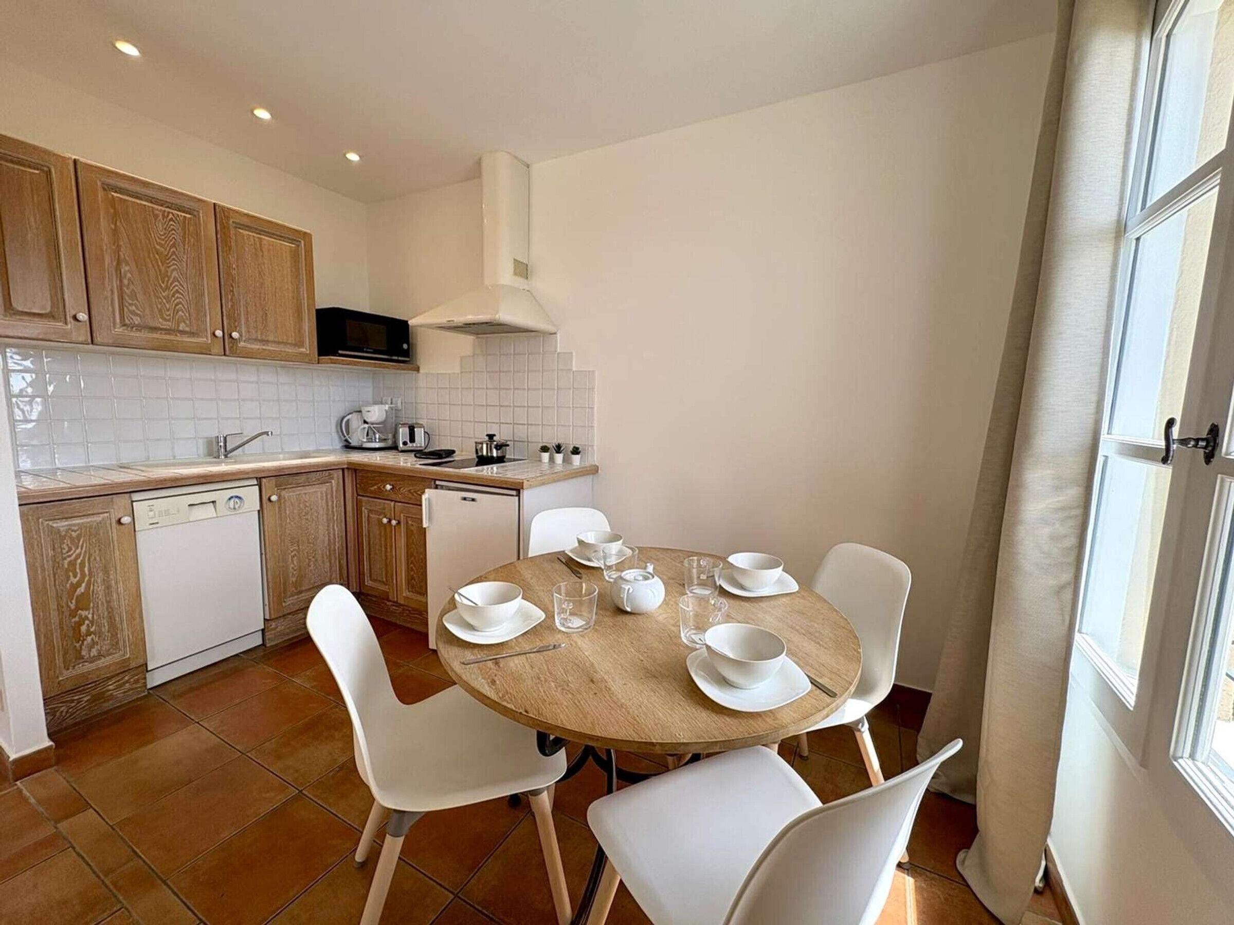 A compact kitchen with white appliances, a dining table with chairs, and a window with curtains.