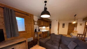 L’intérieur d’un chalet accueillant, avec une table à manger en bois, un canapé gris, un coin cuisine et une fenêtre agrémentée de rideaux.