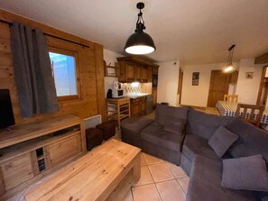 A cozy cabin interior with a wooden dining table, a gray sofa, a kitchen area, and a window with curtains. - Spacious apartment with parking - 8 guests (Les Carroz d'Arâches)