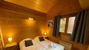 Une chambre dans un chalet en bois, avec deux lits, une fenêtre avec des rideaux, un radiateur et une petite table.