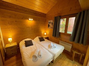 A wooden cabin room with two beds, a window with curtains, a radiator, and a small table. - Spacious apartment with parking - 8 guests (Les Carroz d'Arâches)