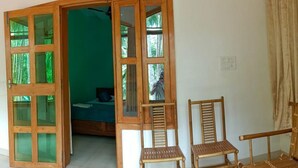 Una habitación con una puerta de madera grande que da a un balcón, una cama con cabecera verde y dos sillas de madera.