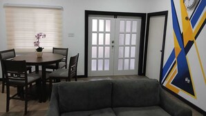 Ein Wohnzimmer mit Sofa, einem Esstisch mit Stühlen, einem Fenster mit Jalousien und einer Tür mit satinierten Glasfeldern.