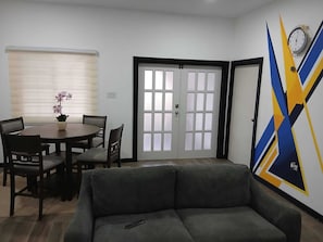 Ein Wohnzimmer mit Sofa, einem Esstisch mit Stühlen, einem Fenster mit Jalousien und einer Tür mit satinierten Glasfeldern.