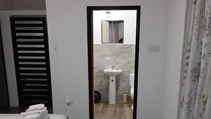 Een smalle gang die leidt naar een badkamer met een wastafel en toilet, een bed met een opgevouwen handdoek, en een deur met een zwart luik.