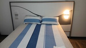 Een bed met gestreepte lakens, een hoofdbord, een bedlampje en een schakelaar aan de muur.