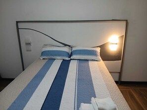 A bed with striped bedding, a headboard, a bedside lamp, and a wall-mounted switch.