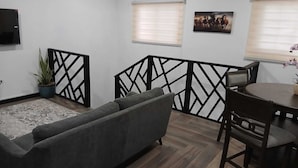 Ein Wohnzimmer mit grauem Sofa, einem Esstisch mit Stühlen, einer Topfpflanze und zwei Fenstern mit Jalousien.