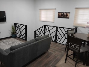 Ein Wohnzimmer mit grauem Sofa, einem Esstisch mit Stühlen, einer Topfpflanze und zwei Fenstern mit Jalousien.