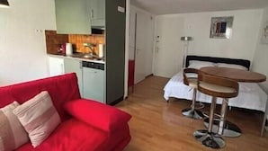 A compact living space with a red sofa, a small kitchen area, a dining table with two stools, and a bed.