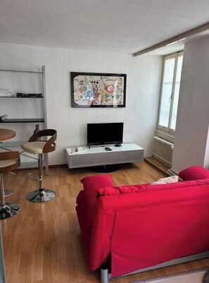Una sala de estar con un sofá rojo, un televisor sobre un soporte blanco, un cuadro en la pared y una ventana.