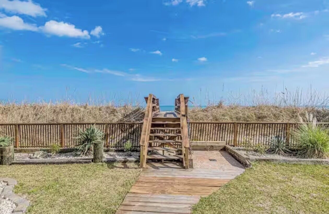 A wooden deck leading to a beach with a wooden fence and sparse vegetation.