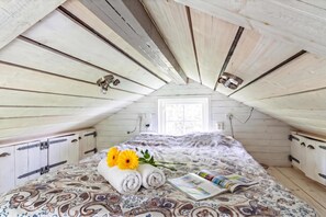 A bedroom with a sloped ceiling, wooden beams, a bed with patterned bedding, a window, and a bedside table with a book and flowers. - The Aspö Hideaway (Drottningskär)