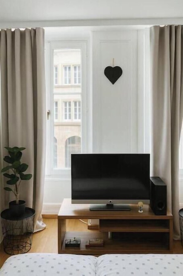 Ein modernes Zimmer mit Fernseher auf einem Holzboden, einem Blumentopf und Blick auf ein Gebäude durch ein Fenster mit Vorhängen.