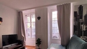 Ein Wohnzimmer mit einer Couch, einem Fernseher auf einem Schrank, einer kleinen Pflanze und Blick auf eine Stadtlandschaft durch das Fenster.