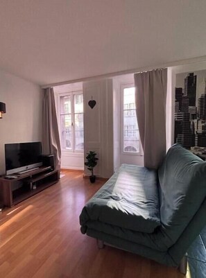 Ein Wohnzimmer mit einer Couch, einem Fernseher auf einem Schrank, einer kleinen Pflanze und Blick auf eine Stadtlandschaft durch das Fenster.