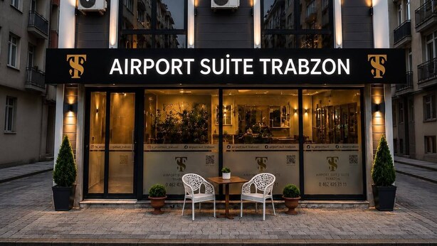 Un edificio con un letrero que dice 'AIRPORT SUITE TRABZON' y un número de teléfono visible. Hay dos plantas en macetas y dos sillas blancas afuera de la entrada.
