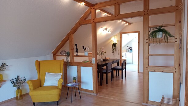 Een kamer met een houten plafond, een gele fauteuil, een eettafel met stoelen, en een keukenruimte zichtbaar door een geopende deur.