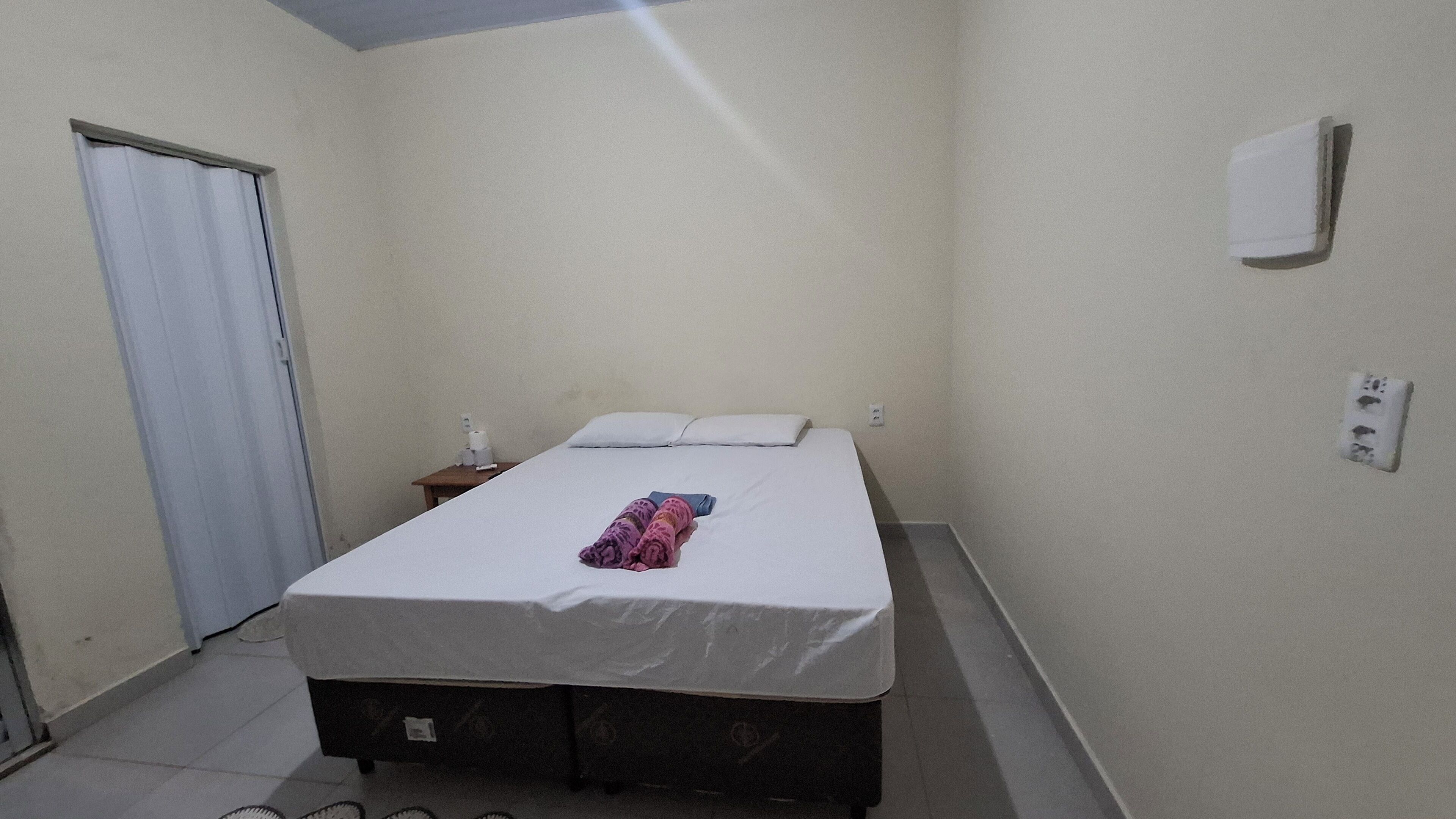 Quarto pequeno com uma cama de solteiro, parede branca lisa, porta fechada, mesa de cabeceira com abajur e uma tomada na parede.
