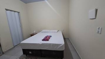 Quarto pequeno com uma cama de solteiro, parede branca lisa, porta fechada, mesa de cabeceira com abajur e uma tomada na parede.