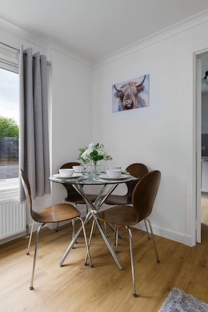Een eetruimte met een glazen tafel, vier stoelen, een vaas met bloemen en een schilderij aan de muur.