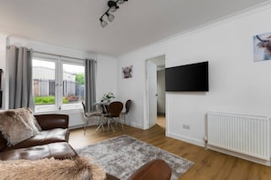 Een moderne woonkamer met een leren bank, een eettafel met stoelen, een aan de muur bevestigde televisie en uitzicht op buiten.