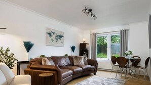 A modern living room with a brown leather sofa, a white armchair, a dining table with chairs, and a large window with curtains. - Forth Court City Retreat (Stirling)