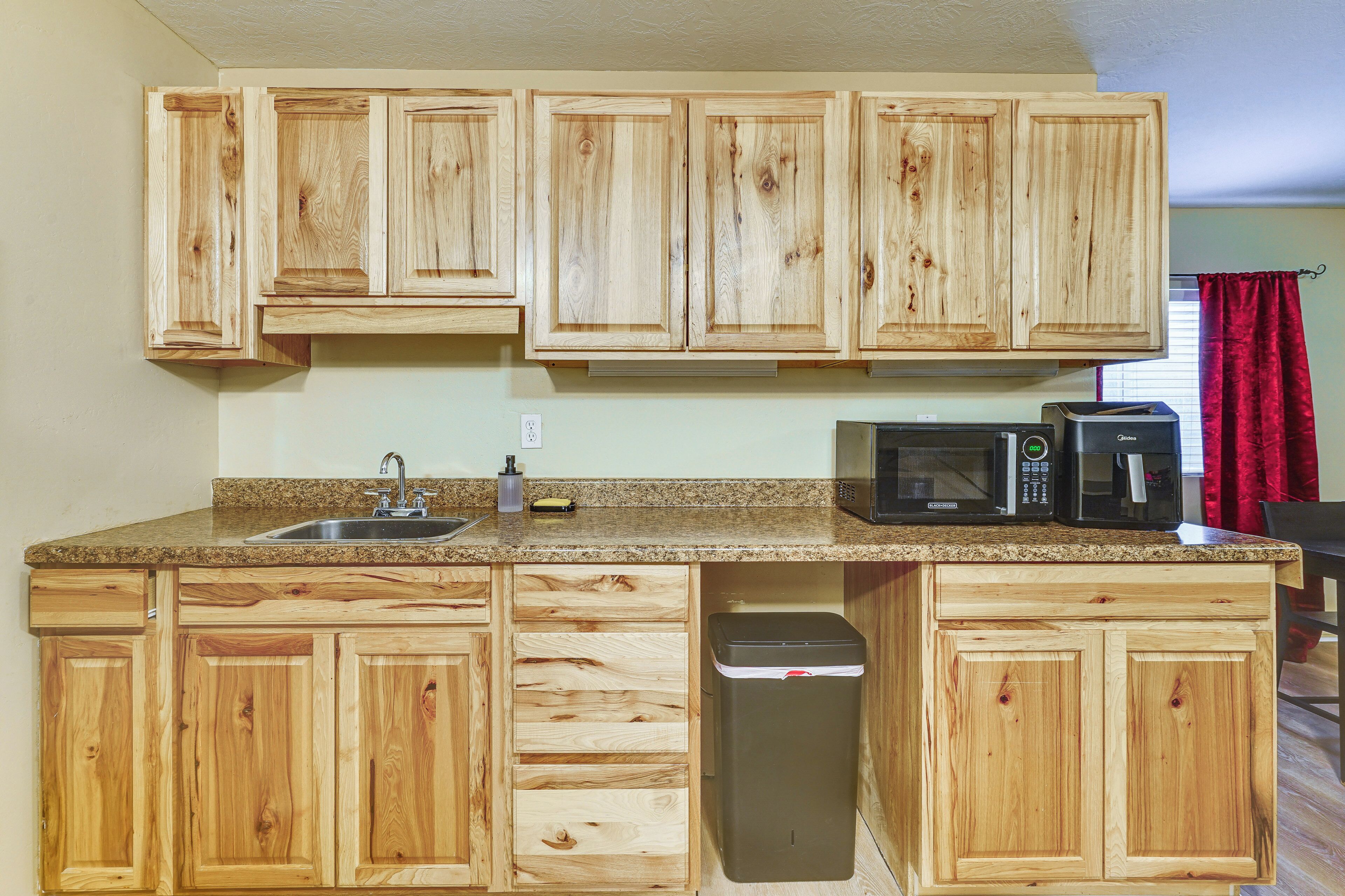 Una cocina con armarios de madera, encimera de granito, fregadero, cubo de basura y microondas.