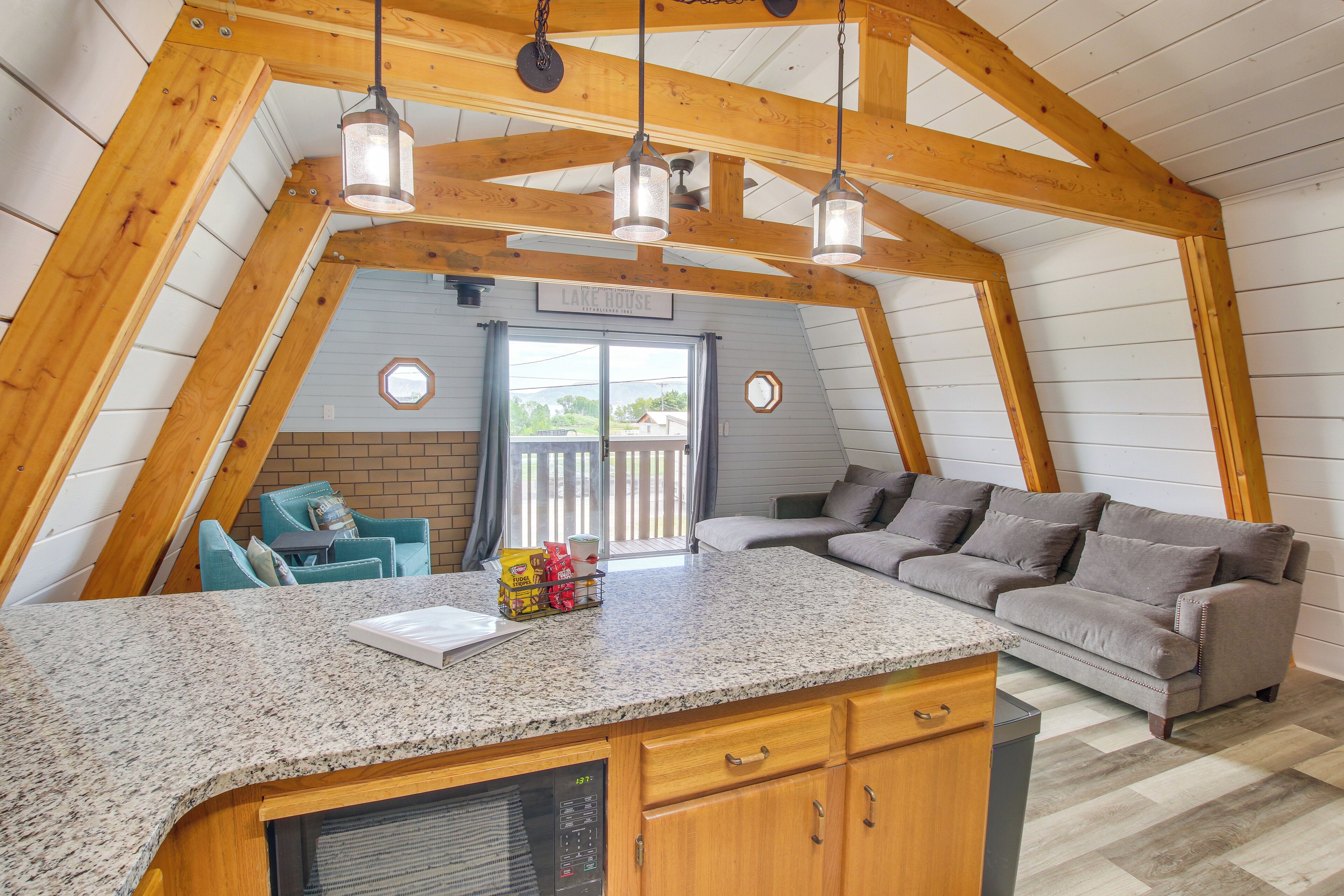 A cozy, triangular room with a kitchen area, a gray sofa, and a window overlooking a deck.