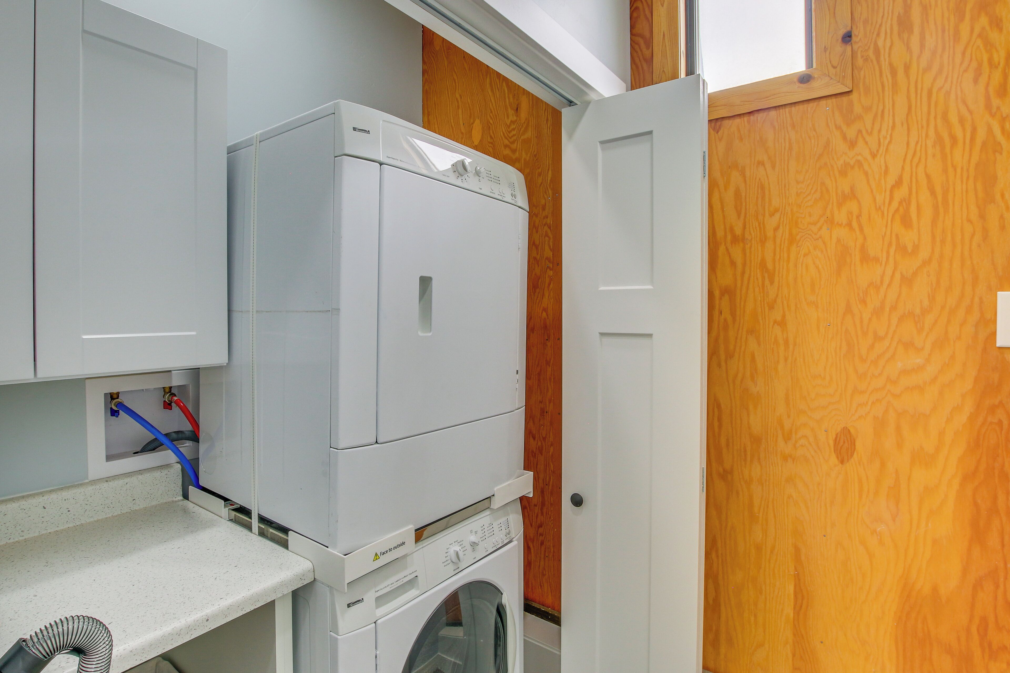 Une buanderie équipée d’un lave-linge et d’un sèche-linge superposés, d  ’une porte en bois et d’un plan de travail blanc avec un tuyau.