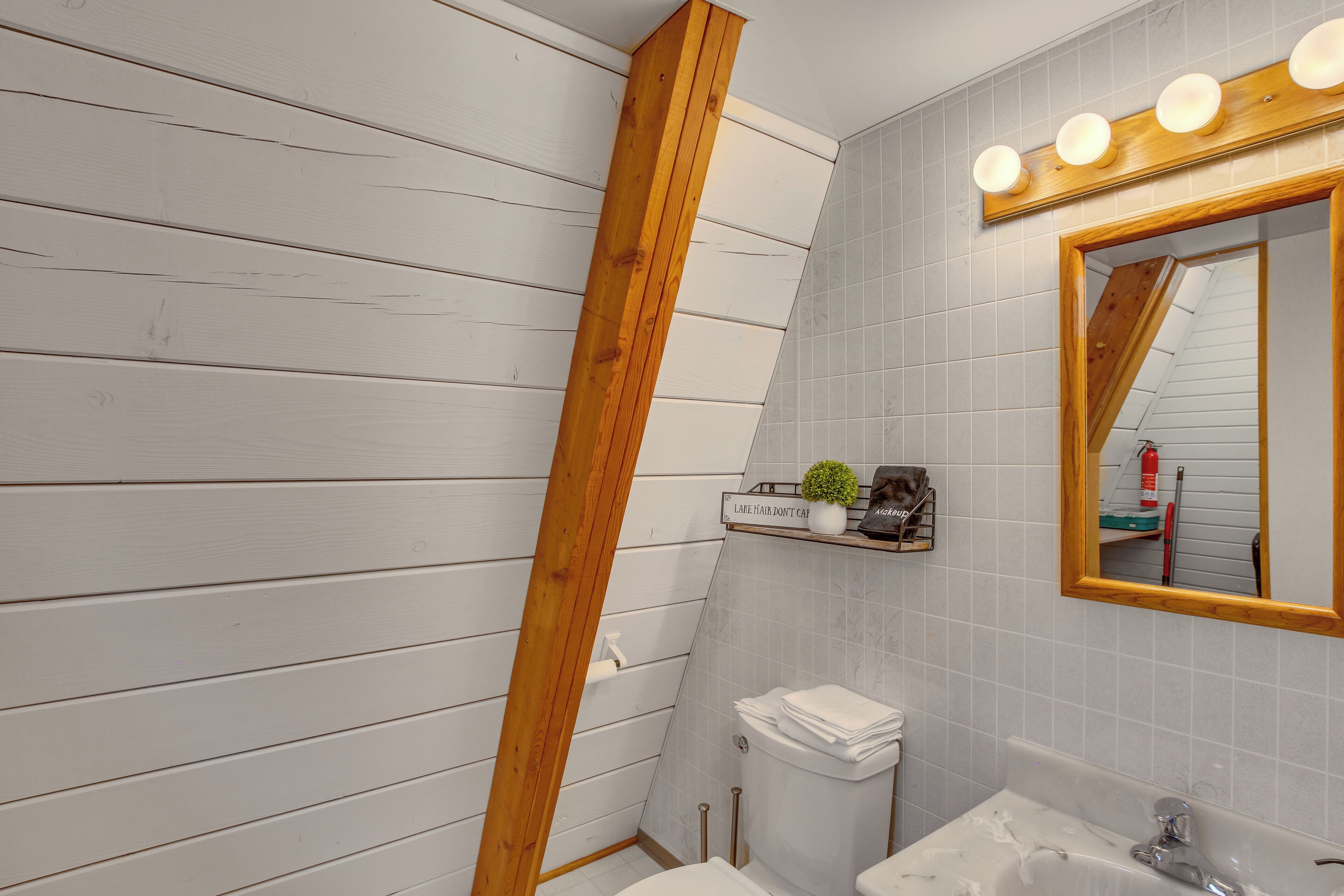 Un baño con una viga de madera, un inodoro, un lavabo, un espejo con iluminación y un estante con artículos de aseo.