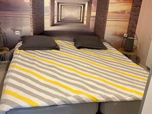Un lit avec une literie à rayures, deux oreillers, une table de chevet avec une lampe et une fresque murale représentant un couloir.